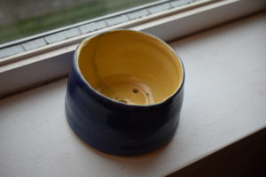Small flower pot. Inside is sunny yellow, outside is dark blue, 4 drainage holes in bottom of pot. Pot is on white windowsill.