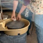 Adding small white clips to hold larger yellow pottery wheel together. Wheel is light yellow and secured on a circular metal base.