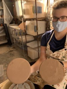 Myself holding two plates. Left plate has been trimmed, edges look very smooth, spiral pattern on bottom of plate. Right plate looks much more rough, no spiral. Some tupperware are visible in the background. 