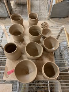 birds eye view of 7 pots and 3 bowls, light brown clay on light brown cork board, various sizes